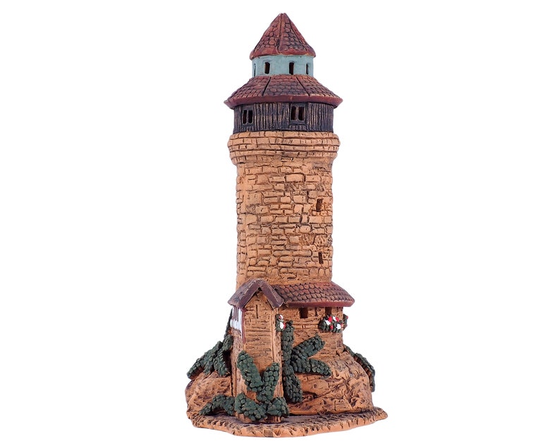May include: A small, brown, ceramic tower with a green roof and a small house attached to the base. The tower has a stone-like texture and is decorated with small windows and a wreath.