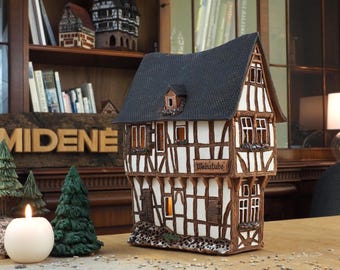 May include: A ceramic model of a traditional German half-timbered house with a black roof, white walls, and brown beams. The house has windows with lights inside and a sign that reads "Weinstube".