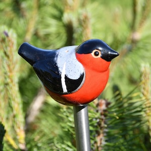 May include: A ceramic bird figurine with black, white, and red paint. The bird is perched on a metal stake, which is inserted into the ground.