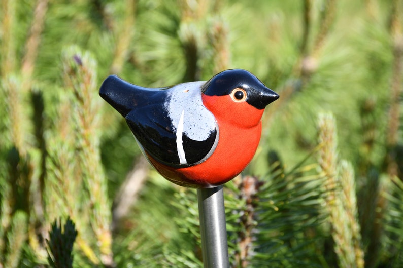 May include: A ceramic bird figurine with black, white, and red paint. The bird is perched on a metal stake, which is inserted into the ground.