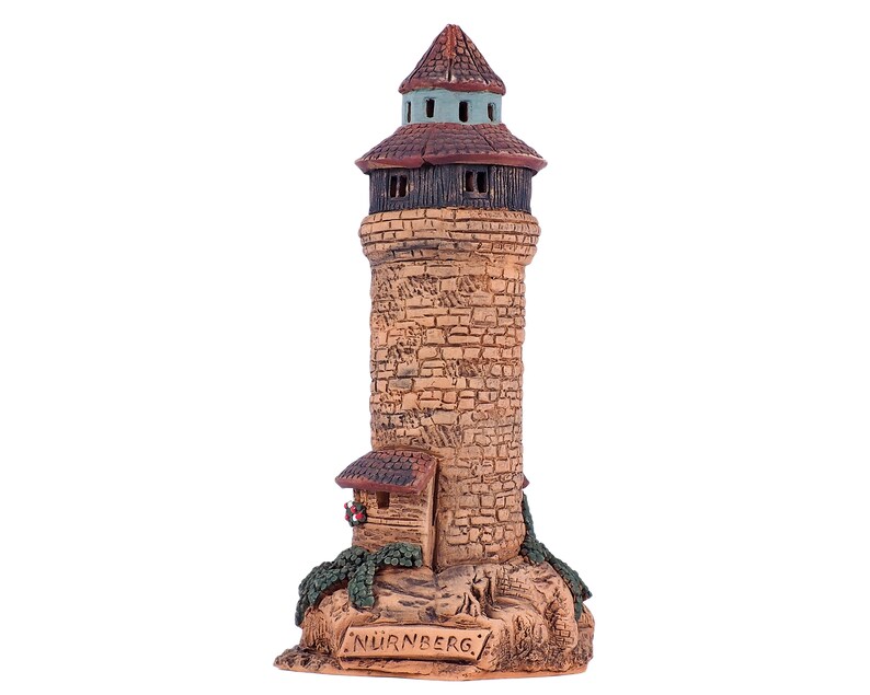 May include: A ceramic model of a tower with a brown roof and a small house at the base. The tower is made to look like stone and has a sign that reads "NURNBERG".