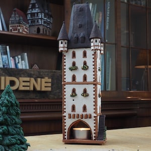 May include: A white ceramic tower with a black roof and a small candle inside. The tower has a decorative design with windows and balconies. The tower is sitting on a wooden surface.