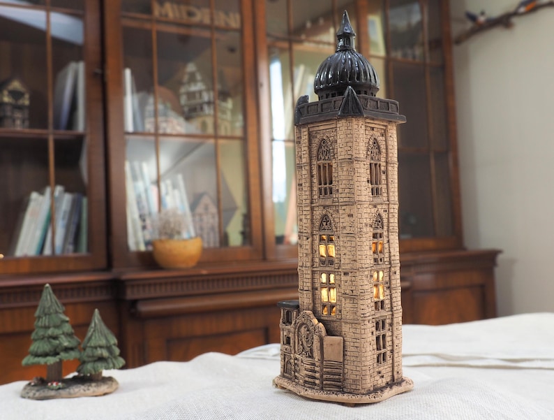 May include: A detailed, beige ceramic tower with a black dome, featuring multiple windows that emit a warm, yellow glow. The tower is designed with architectural details, resembling a historic building. Two small green decorative pine trees are in the foreground.