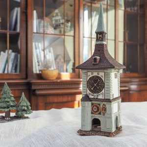 May include: A detailed ceramic clock tower figurine with a brown roof, a clock face, and a tall spire. The tower is painted in shades of green, brown, and white. Two small green Christmas tree figurines are in the foreground.