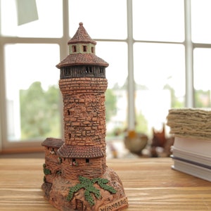 May include: A small, detailed ceramic model of a tower with a brown roof and brick walls. The tower is surrounded by green foliage and has the word "N&Uuml;RNBERG" written on the base.