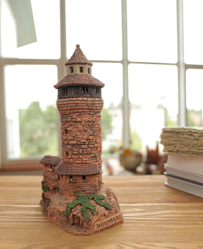 May include: A small, detailed ceramic model of a tower with a brown roof and brick walls. The tower is surrounded by green foliage and has the word "N&Uuml;RNBERG" written on the base.