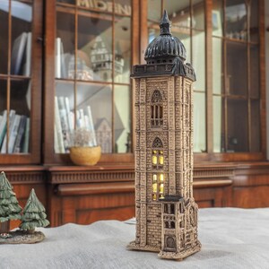 May include: A detailed, beige-colored ceramic tower with a black dome and arched windows. The tower is illuminated from within, suggesting a lamp or decorative piece. Two small green ceramic Christmas trees are in the foreground.