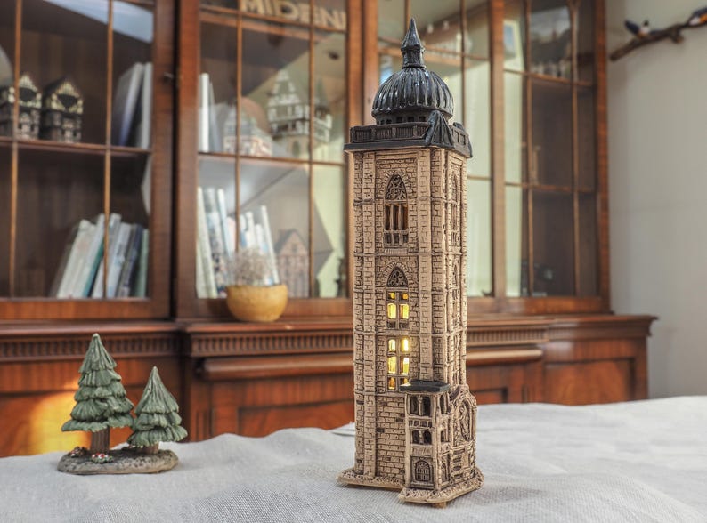 May include: A detailed, beige-colored ceramic tower with a black dome and arched windows. The tower is illuminated from within, suggesting a lamp or decorative piece. Two small green ceramic Christmas trees are in the foreground.