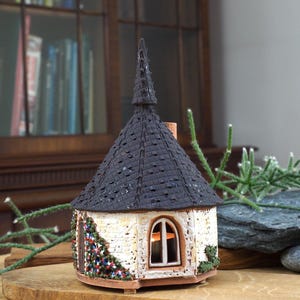 Puede incluir: Una pequeña cabaña de cerámica blanca con un techo de tejas negras y una aguja puntiaguda. La cabaña tiene una sola ventana con una vela en el interior y está decorada con hiedra verde y roja.