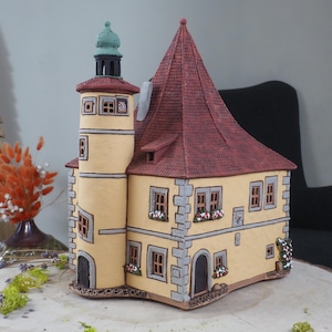 May include: A hand-painted ceramic model of a yellow building with a red roof and a tower. The building has windows and a door, and there are flowers on the windowsills.