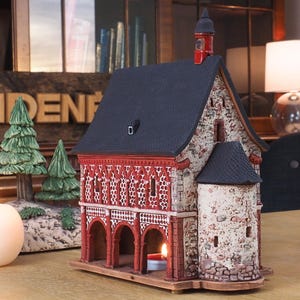 May include: A ceramic model of a small church with a red brick facade, a black roof, and a white tower. The model is on a wooden base and has a small candle inside.