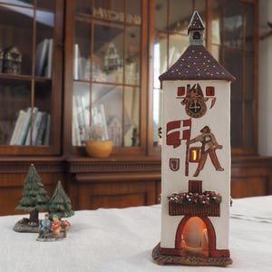 May include: A white ceramic tower with a red roof and a lit candle inside. The tower has a painted design of a person holding a flag and a flower box with red flowers. The tower is on a white tablecloth.