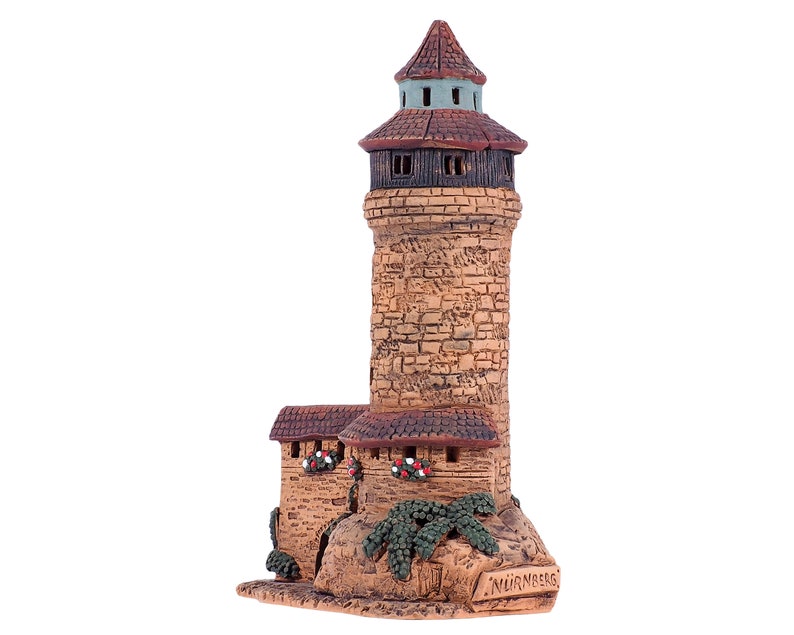 May include: A small ceramic model of a tower with a brown roof and stone walls. The tower is decorated with green plants and the word "NURNBERG" is written on the base.