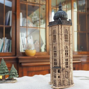 May include: A detailed, beige ceramic tower with a dark dome and intricate window designs. The tower is a decorative piece, possibly a model of a historical building. Two small green tree figurines are in the foreground.