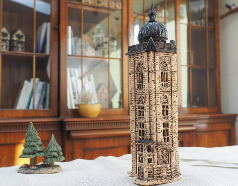 May include: A detailed, beige ceramic tower with a dark dome and intricate window designs. The tower is a decorative piece, possibly a model of a historical building. Two small green tree figurines are in the foreground.