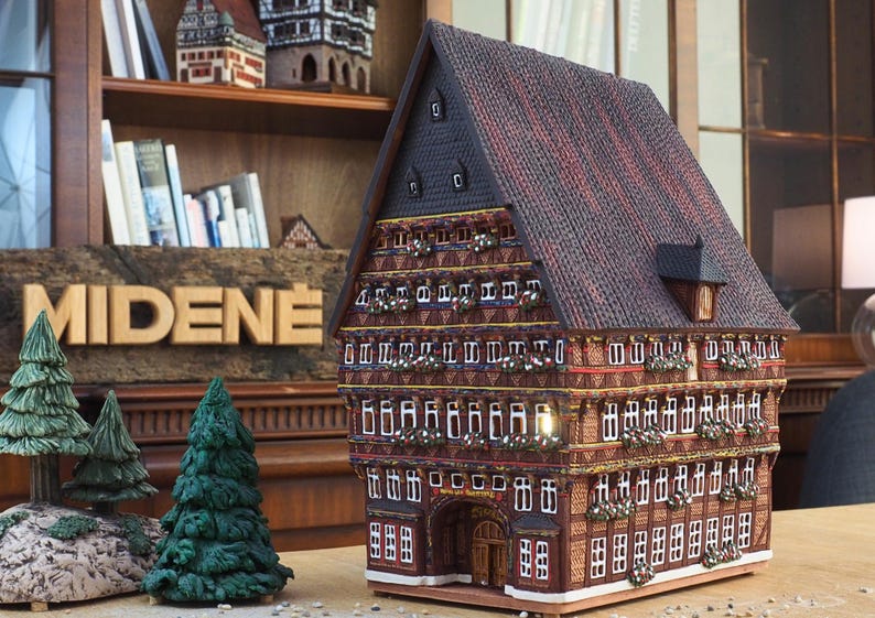 Puede incluir: Un modelo detallado de una casa tradicional alemana de entramado de madera con un techo de tejas rojas y muchas ventanas. La casa est&aacute; decorada con detalles intrincados y tiene un letrero que dice "MIDENE".