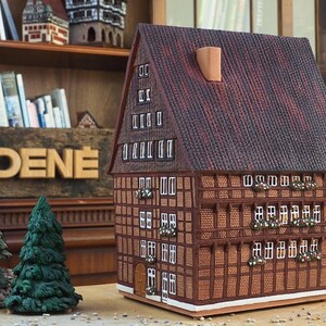 Puede incluir: Un modelo detallado de una casa tradicional alemana de entramado de madera con un techo de tejas marrones y molduras blancas. La casa tiene varias ventanas y una peque&ntilde;a puerta. El modelo est&aacute; hecho de madera y est&aacute; sentado sobre una superficie de madera.