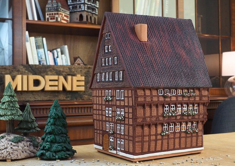 Puede incluir: Un modelo detallado de una casa tradicional alemana de entramado de madera con un techo de tejas marrones y molduras blancas. La casa tiene varias ventanas y una peque&ntilde;a puerta. El modelo est&aacute; hecho de madera y est&aacute; sentado sobre una superficie de madera.