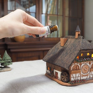 Peut inclure: Une maison en céramique en forme de chalet avec un toit de chaume et une roue à eau fonctionnelle. La maison possède un cadran d'horloge et une cheminée. Une main tient une petite bouteille d'huile essentielle au-dessus de la cheminée.