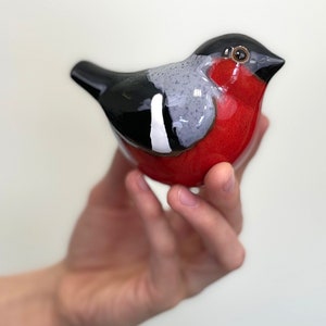 May include: A ceramic bird figurine with a red, black, and gray painted design. The bird is sitting in a hand.