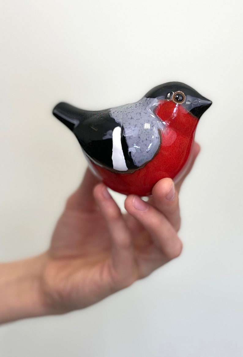 May include: A ceramic bird figurine with a red, black, and gray painted design. The bird is sitting in a hand.