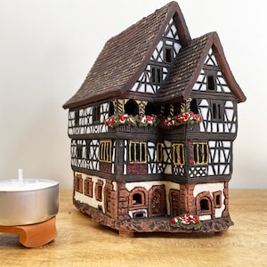 Könnte beinhalten: Eine Keramik-Hausfigur mit braunem Dach, weißen Wänden und schwarzen Verzierungen. Das Haus hat mehrere Fenster und Balkone mit roten Blumen. Die Figur steht auf einer Holzoberfläche.