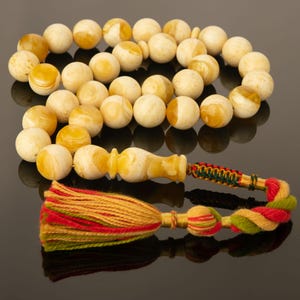 May include: A string of prayer beads featuring smooth, round, marbled cream and honey-coloured amber-like spheres. The strand includes a larger, elongated central bead and terminates in a decorative, multi-coloured tassel with red, green, and yellow threads.