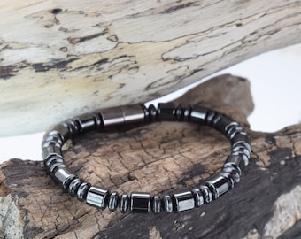 Op de afbeelding: Een donkergrijze magnetische armband bestaande uit kleine, vierkante en cilindrische kralen. De armband wordt getoond op een stuk verweerd hout, met een witte achtergrond. De armband heeft een magnetische sluiting.