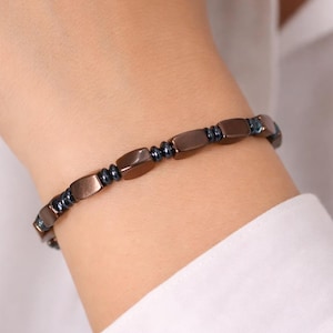 Könnte beinhalten: Ein kupferfarbenes Armband mit rechteckigen Perlen und kleineren, dunkelblauen Perlen. Das Armband wird am Handgelenk getragen und hat ein schlichtes, elegantes Design. Das Armband ist ein Schmuckstück.