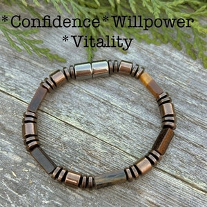 Könnte beinhalten: Ein braunes und goldenes Perlenarmband mit einem silbernen Magnetverschluss. Das Armband trägt die Worte "Selbstvertrauen", "Willenskraft" und "Vitalität" auf dem Hintergrund.