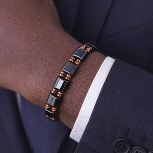 May include: A black and copper-colored bracelet with rectangular links is worn on a wrist. The bracelet is made of black rectangular links with copper-colored accents. The person is wearing a navy blue suit with a white dress shirt.