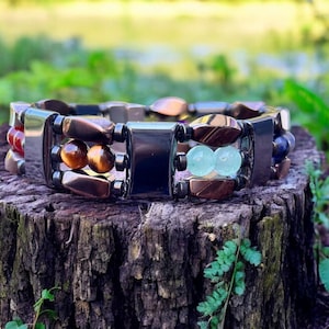 May include: A multi-coloured beaded bracelet with brown, black, red, and green stones. The bracelet is made of metal and has a magnetic clasp.
