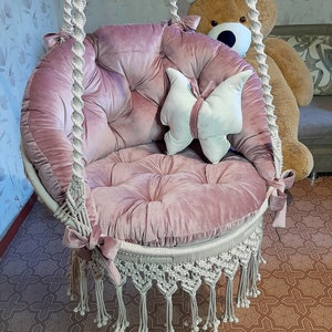 K&ouml;nnte beinhalten: Ein runder, rosa Samt-Schaukelstuhl mit Knopfheften und Makramee-Details. Der Stuhl hat einen wei&szlig;en Makramee-Fransenbesatz und ist an einem dicken, gedrehten Seil aufgeh&auml;ngt. Ein schmetterlingsf&ouml;rmiges Kissen liegt im Inneren.