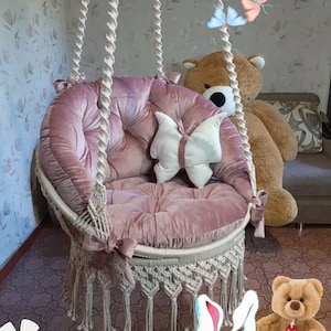 Peut inclure: Un fauteuil suspendu en velours rose avec des détails en macramé et un coussin en forme de papillon. La chaise est suspendue par de grosses cordes tressées. Un grand ours en peluche est derrière la chaise.