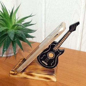 May include: A wooden phone stand featuring a carved guitar design. The stand is made of light brown wood with a dark brown stain. The guitar is black with white strings.