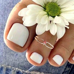 May include: A close-up of a foot with white nail polish and a toe ring with a pink gem. A white daisy flower rests on the foot. The image is set against a denim background.