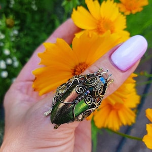 May include: A handcrafted ring with a beetle design. The ring features two iridescent green beetle wings, intricately wrapped in bronze-coloured wire. Small blue and purple beads add accents. The ring is held near bright yellow flowers.