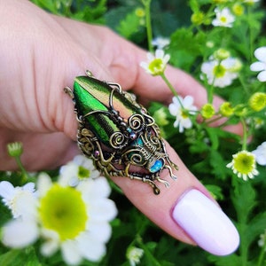 May include: A unique, handcrafted ring featuring a large, iridescent green beetle wing set in an ornate, antique brass-toned metal frame. The ring has intricate wirework and small, colorful accents, creating a statement piece. The ring is shown against a backdrop of green foliage and white flowers.