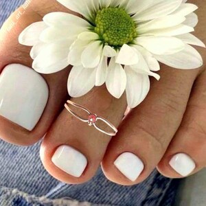 May include: Close-up of a foot with white nail polish and a toe ring with a pink gem. A white daisy flower rests on the foot. The image is set against a denim background.