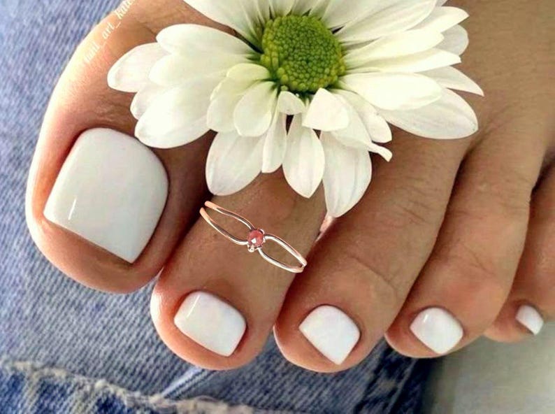 May include: Close-up of a foot with white nail polish and a toe ring with a pink gem. A white daisy flower rests on the foot. The image is set against a denim background.