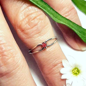 May include: A silver ring with a small, round, red gemstone is worn on a finger. The ring has a delicate, split-shank design. Green leaves and white flowers with yellow centers are in the background.