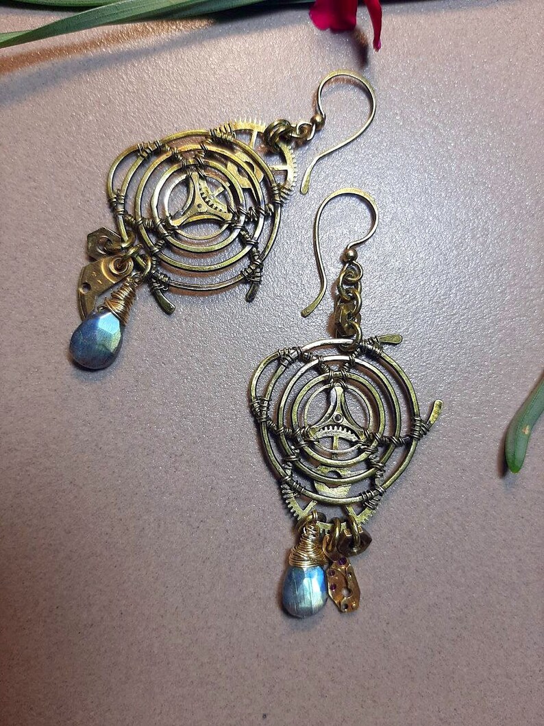 Może przedstawiać: Para złotych kolczyk&oacute;w w stylu steampunk. Każdy kolczyk ma okrągły kształt z kołami zębatymi i niebieskim kamieniem szlachetnym w kształcie łzy.