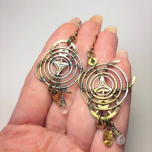 Może przedstawiać: Para kolczyk&oacute;w steampunk w kolorze złotym i srebrnym o okrągłym wzorze z k&oacute;ł zębatych i drutu. Każdy kolczyk ma mały wisiorek z kryształu.