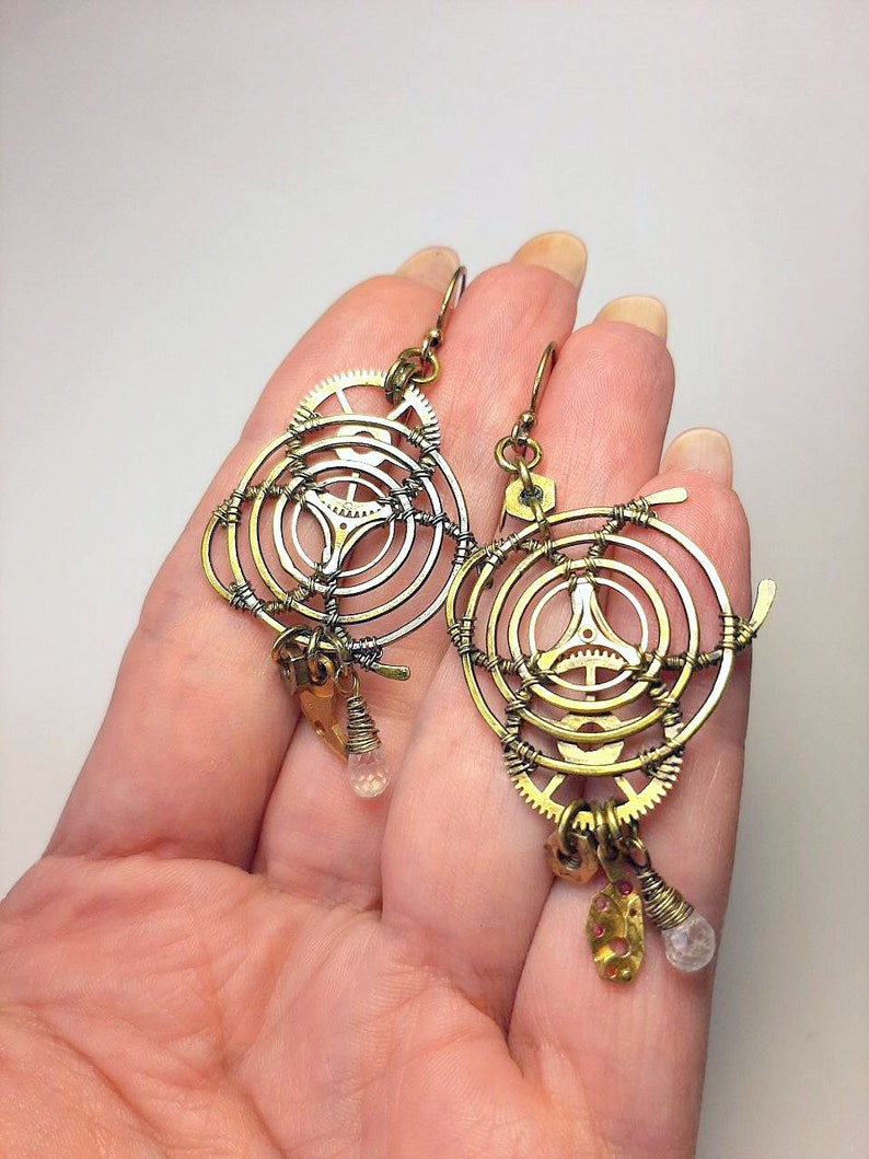 Może przedstawiać: Para kolczyk&oacute;w steampunk w kolorze złotym i srebrnym o okrągłym wzorze z k&oacute;ł zębatych i drutu. Każdy kolczyk ma mały wisiorek z kryształu.