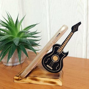 May include: A wooden phone stand featuring a black guitar design. The stand is crafted from natural wood with a burnt finish. The guitar is a simple silhouette with a few details.