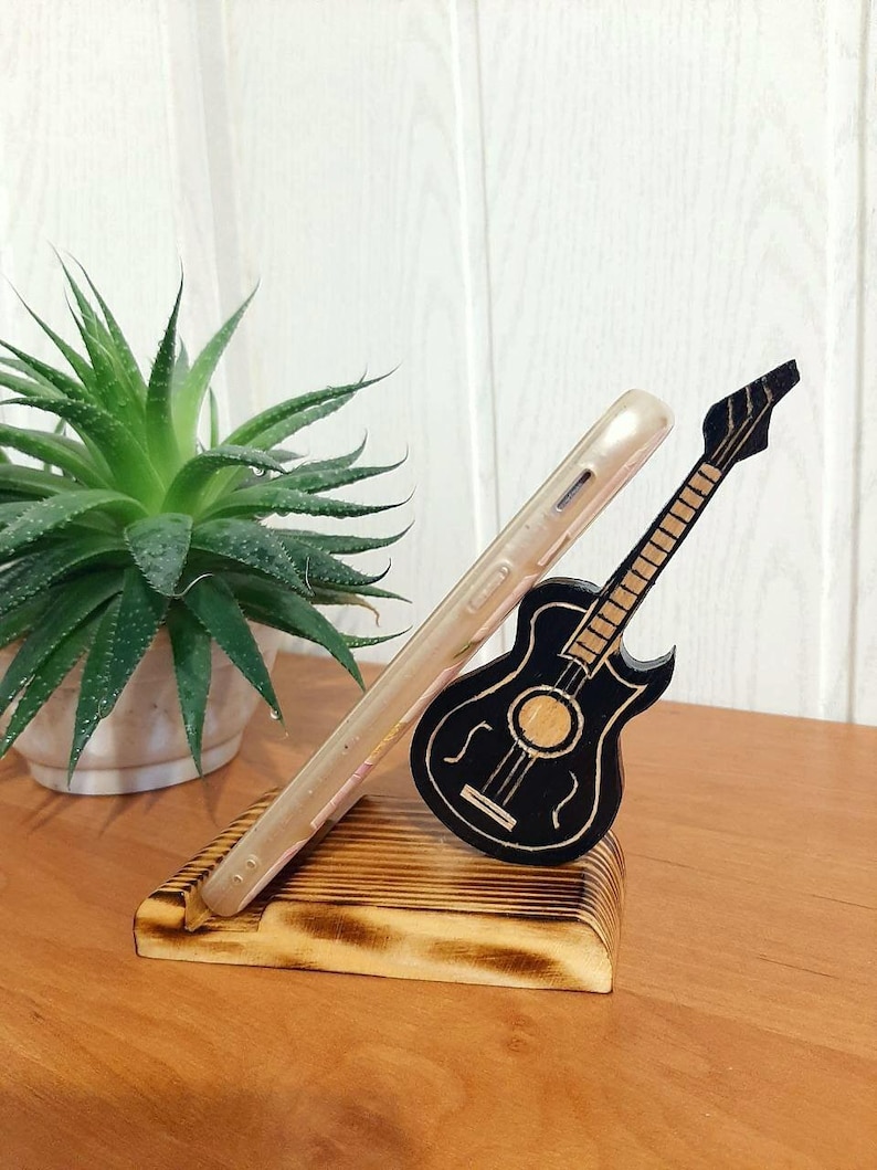 May include: A wooden phone stand featuring a black guitar design. The stand is crafted from natural wood with a burnt finish. The guitar is a simple silhouette with a few details.