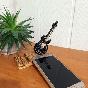 May include: A wooden phone stand with a black guitar design. The stand is holding a gold smartphone with a black screen.