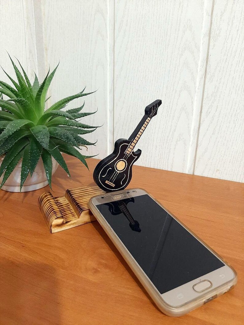 May include: A wooden phone stand with a black guitar design. The stand is holding a gold smartphone with a black screen.