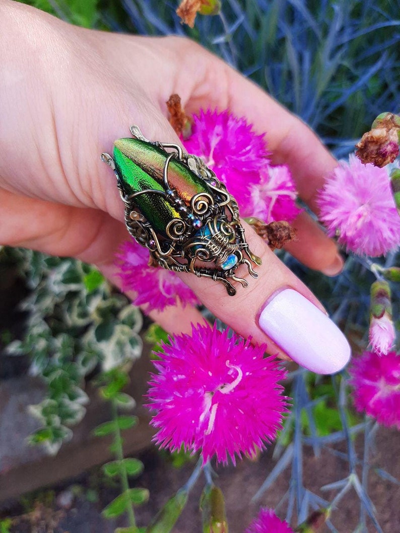 May include: A unique ring featuring a large, iridescent green beetle wing set in an ornate, wire-wrapped bronze setting. The ring has intricate details with small, colorful accents. The background includes pink flowers and greenery.