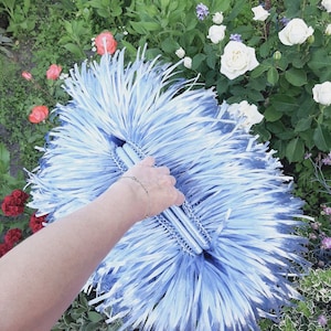 May include: A light blue clutch bag featuring a dense, shaggy texture made of numerous thin, ribbon-like strips. The bag has a central horizontal opening and a rounded, elongated shape, with the light blue material radiating outward from the center.